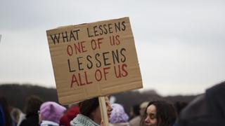 woman holding sign "what lessens one of us lessens all of us"