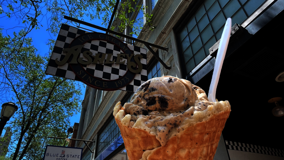 A photograph of a scoop of Coffee Oreo ice cream in a waffle cone in front of the sign for Ashely's Ice Cream