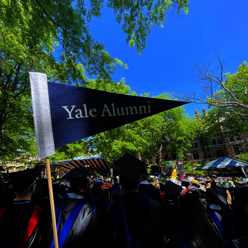 A Yale Alumni pennant stands at the foreground, with graduates from the Yale Class of 2022 behind it.